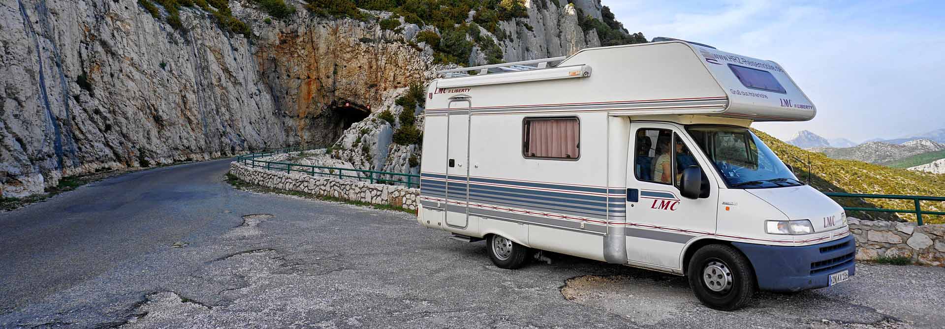A white camper van with blue and red stripes is parked on a scenic mountain road, perfect for exploring or climate-controlled camper storage. In the background, a rocky cliff wall with greenery crowns the entrance to a mysterious tunnel.