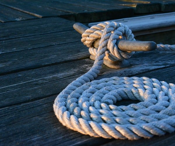 Close-up of a coiled rope tied securely to a metal cleat on a wooden dock, reminiscent of indoor boat storage. The neatly arranged rope rests over weathered wood, with soft lighting casting gentle shadows across the scene.