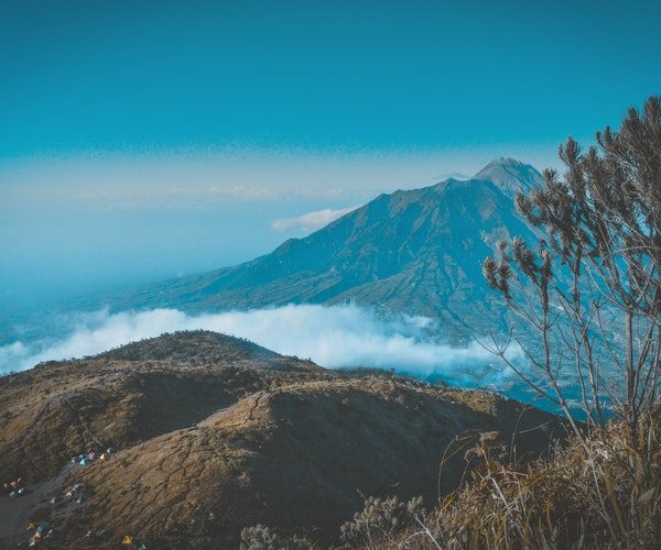A scenic view of a mountainous landscape with a prominent peak under a clear blue sky. Low clouds hover around the mountain base. In the foreground, shrubs and grasses surround small colorful tents on the hillside, evoking a sense of tranquility often sought in indoor RV storage escapes.