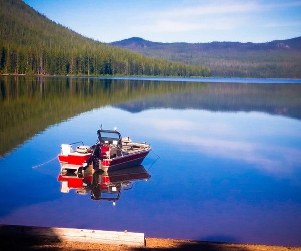 A red and white boat is anchored on a serene, mirror-like lake reflecting trees and a blue sky. A person in a red jacket stands on the boat, surrounded by calm water and distant greenery, perfectly situated for boat storage when not exploring the tranquil surroundings.
