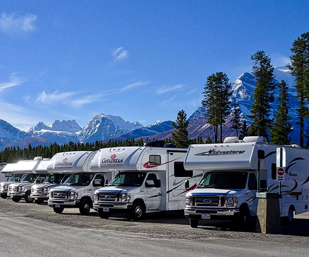 A row of parked white recreational vehicles (RVs) with branding, set against a backdrop of snow-capped mountains and a forested area under a clear blue sky, highlights the contrast between their rugged beauty and the practicality of indoor camper storage when its time to protect them from the elements.