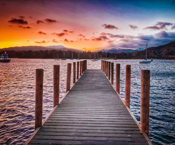 A wooden dock extends into a tranquil lake with sailboats and a ferry in the distance. The water reflects the vibrant colors of a sunset, while climate-controlled boat storage facilities ensure your vessels safekeeping as mountains rise on the horizon under a partly cloudy sky.