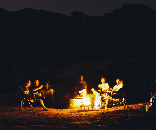Five people sit around a glowing campfire at night, relaxed in folding chairs and engaged in conversation. The fire casts a warm light, illuminating their faces and creating a cozy outdoor atmosphere that starkly contrasts with memories of climate-controlled camper storage back home.