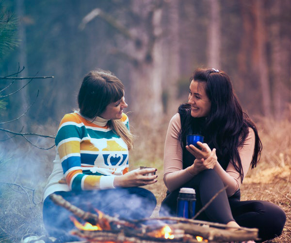 Two people are sitting outdoors, smiling and holding cups. One wears a colorful striped sweater, and the other wears a light pink top near their camper storage. They are surrounded by blurred natural scenery, suggesting a relaxed, friendly atmosphere.