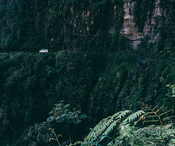 A narrow winding road runs along a steep, lush green cliffside. A single white vehicle navigates the path, reminiscent of RV storage tales amidst dense vegetation and towering cliff walls. The scene evokes a sense of adventure and remote wilderness.