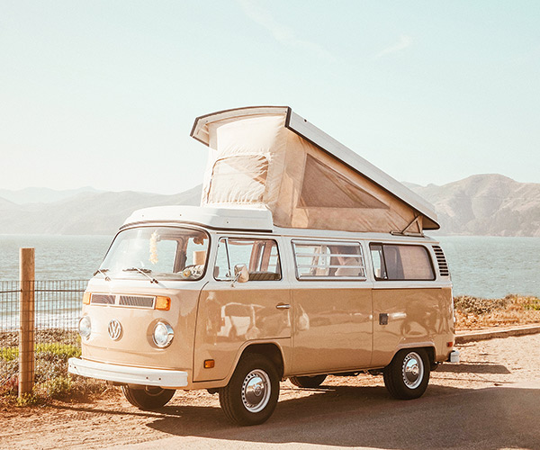 A vintage beige Volkswagen camper van with an open pop-top roof is parked by a coastal road, offering a taste of adventure before retreating to RV storage. The ocean and mountains provide a stunning backdrop under the clear sky.