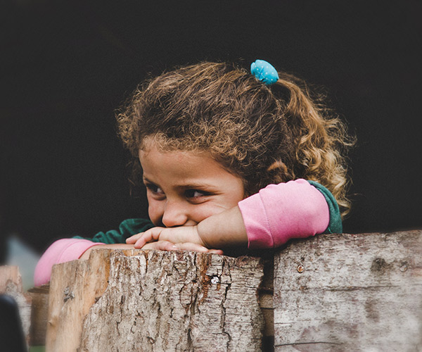 A young child with curly hair and a blue hair tie leans on a rustic wooden fence, gazing thoughtfully into the distance. The background is dark, like the quiet interior of climate controlled RV storage, highlighting the childs contemplative expression.