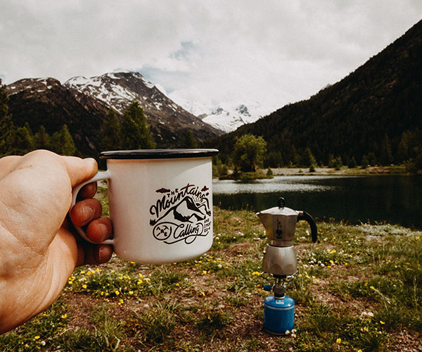 A hand holds a mug adorned with mountain graphics, capturing the essence of adventure. In the backdrop, a scenic vista features mountains, a lake, and a coffee pot on a portable stove—a testament to life beyond indoor camper storage amid lush greenery and snow-capped peaks under a cloudy sky.